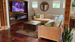 A cozy living room with a wooden ceiling, a large flat-screen TV, a beige sofa, a wicker chair, a coffee table, and a small side table with a lamp.