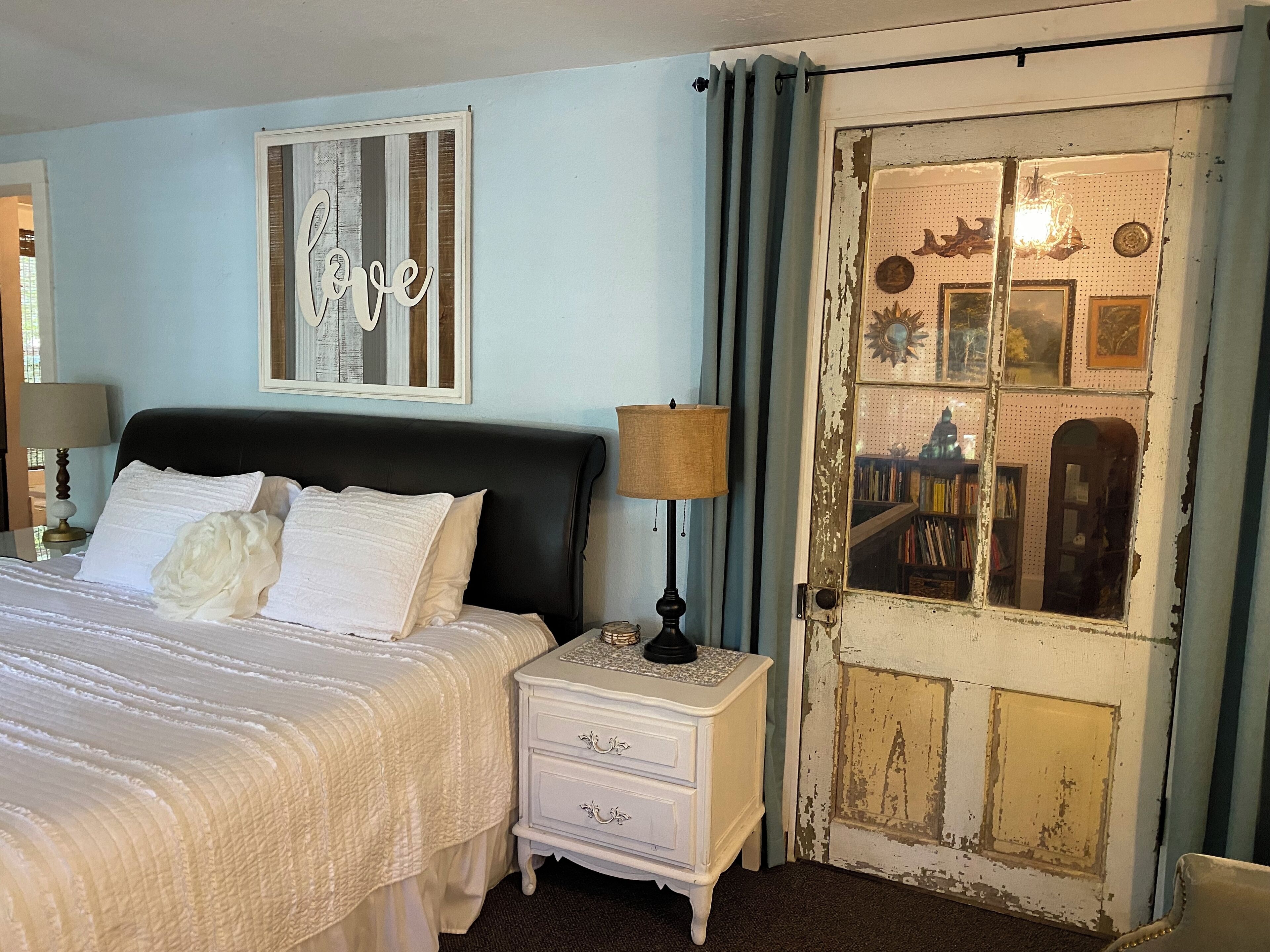 Une chambre avec un lit, une table de chevet, une commode, un mur orné d’un panneau avec le mot « amour », et une porte au style rétro.