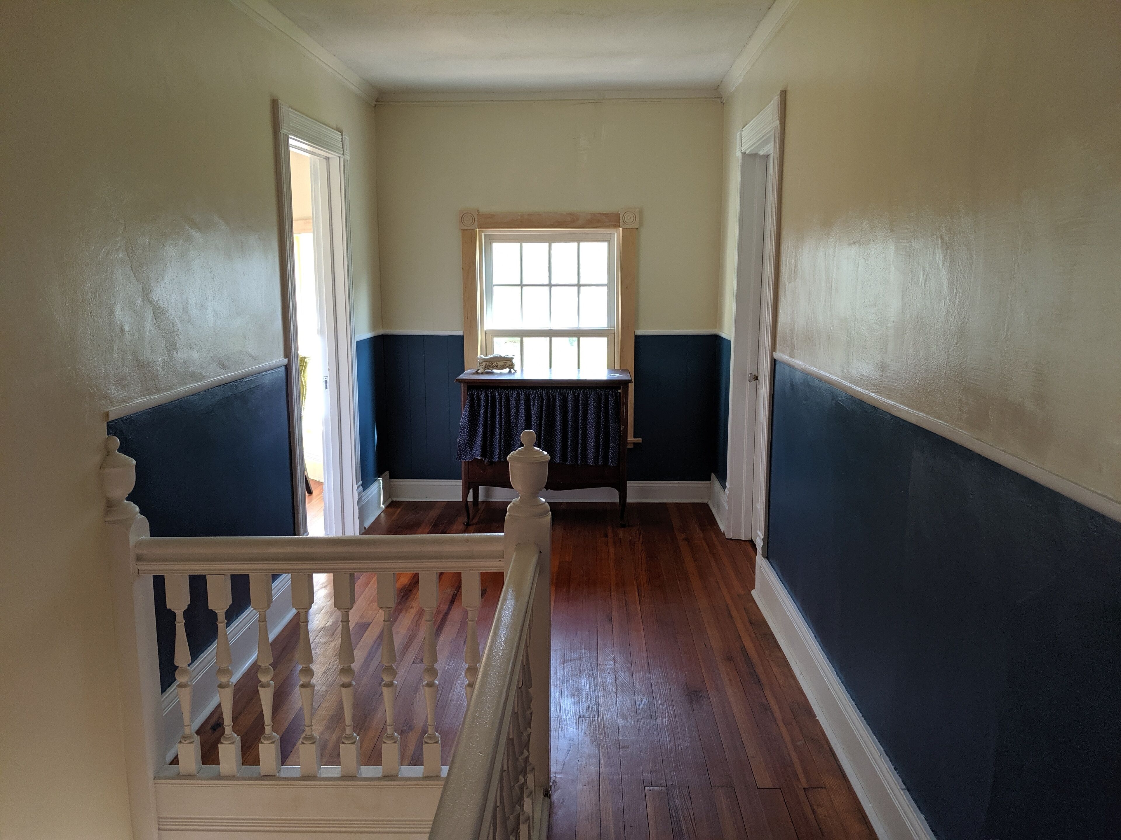 A hallway with wooden flooring, a staircase with white balusters, and a window with a table and chair in the background.