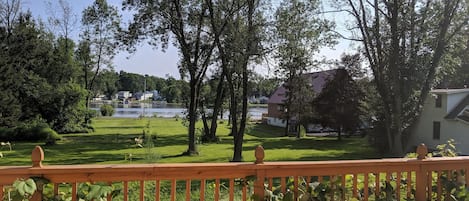 A view from a balcony with a wooden railing, overlooking a grassy area with trees and a body of water in the background.