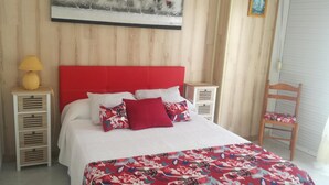 A bedroom with a red headboard, white bedding, and a floral-patterned bedspread. There is a wooden nightstand with a lamp and a chair with a patterned cushion.
