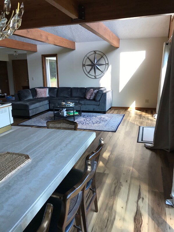 Un salon moderne avec un canapé d’angle, une table à manger avec des chaises et une grande fenêtre laissant entrer la lumière naturelle.