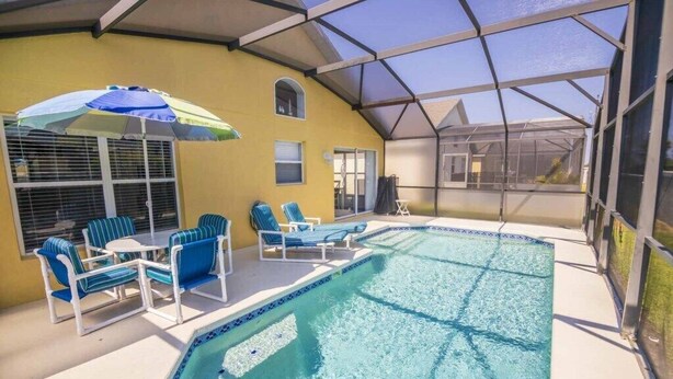 Una piscina techada con sillas de descanso y una sombrilla, rodeada por un edificio amarillo con cerramiento de vidrio.