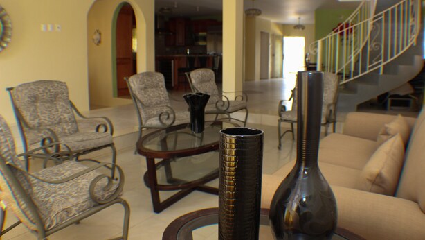 A living room with a sofa, two patterned armchairs, a glass coffee table, and a tall vase. A staircase with a metal railing is visible in the background.