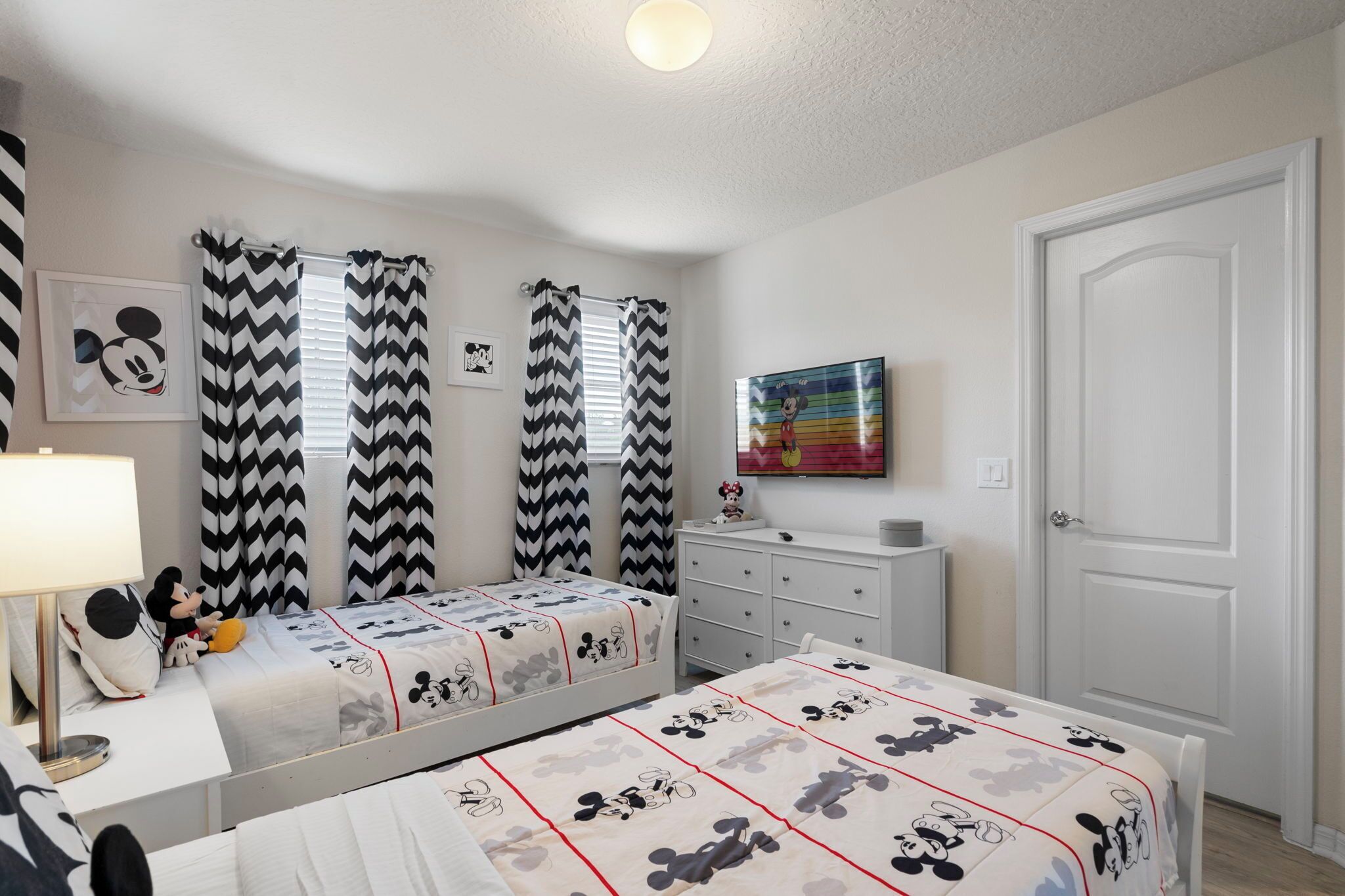 Une chambre avec deux lits, chacun recouvert d’une parure de lit aux motifs de Mickey Mouse. On y trouve une commode, un miroir et un doudou à l’effigie de Mickey Mouse sur l’un des lits. La chambre a une porte blanche et un téléviseur fixé au mur.