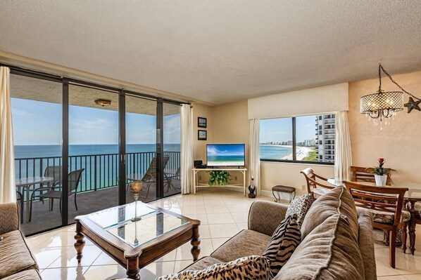 A spacious living room with a glass coffee table, a sofa with leopard print cushions, a television on a stand, and a balcony with a view of the ocean. - Enjoy Paradise in this 2/2 Beachfront Condo (Marco Island)