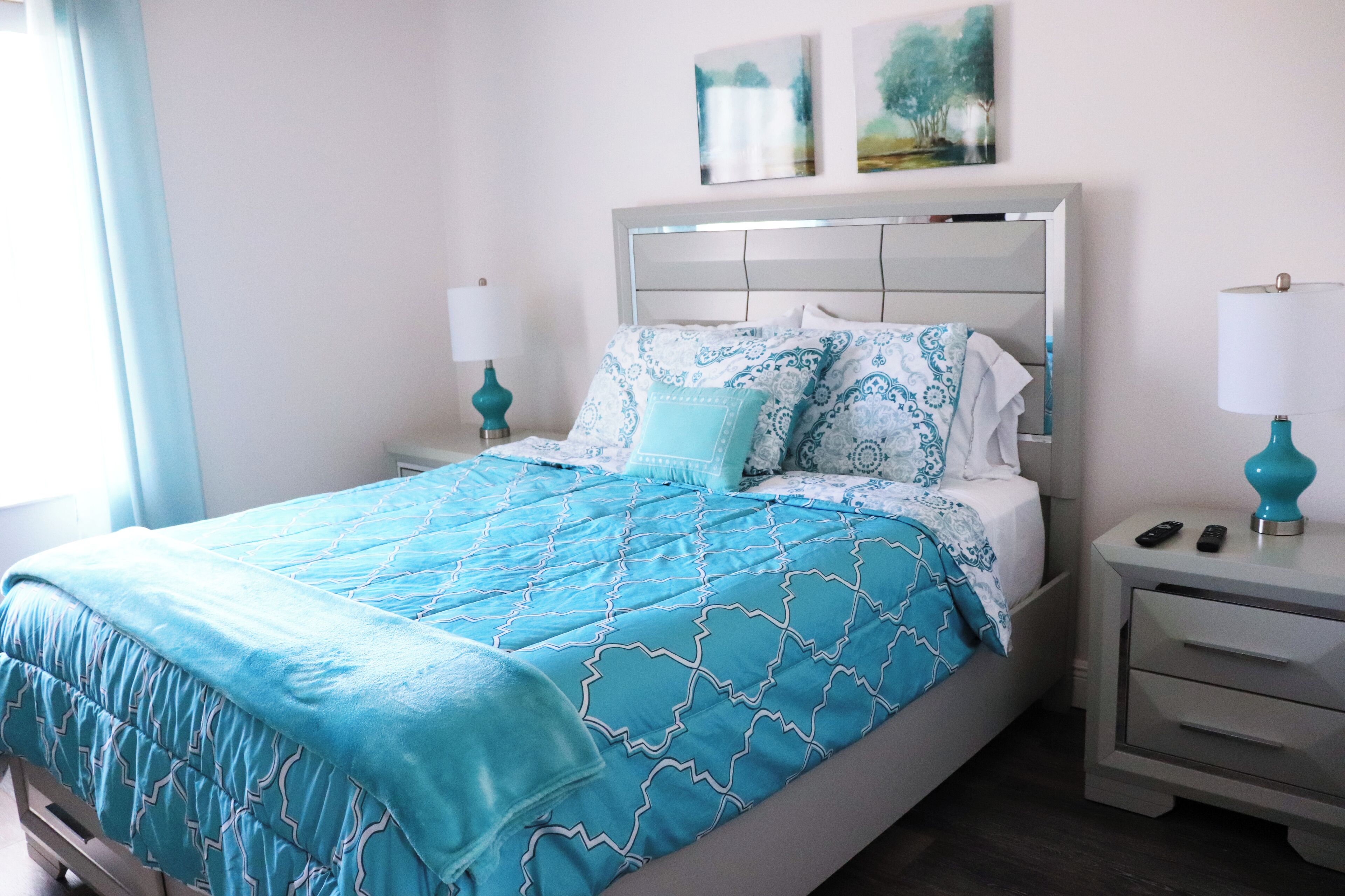 Una cama bien hecha con una colcha azul y blanca estampada, dos lámparas de noche y una mesita de noche con un control remoto.