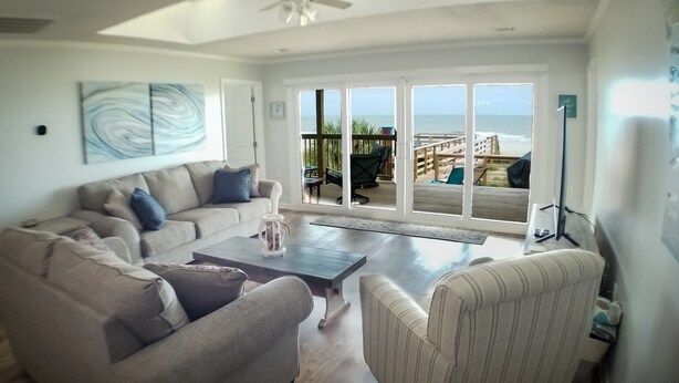 A living room with a sofa, armchair, coffee table, and a view of the beach through large sliding glass doors.