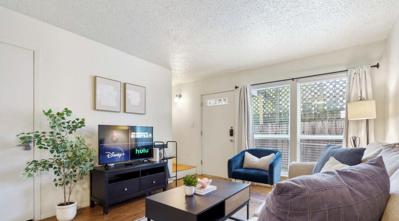 Een moderne woonkamer met een flatscreen televisie, een plant in pot, een bank, een salontafel en een glazen schuifdeur naar een balkon.