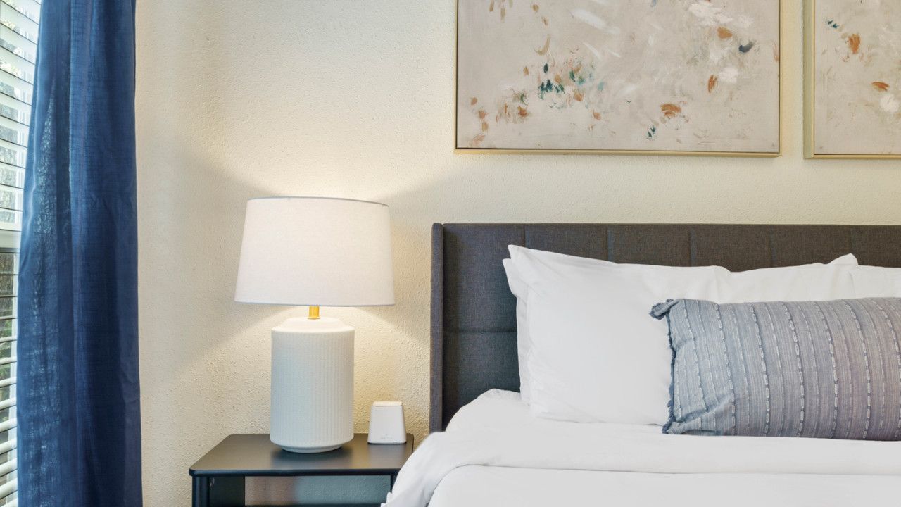 Een net opgemaakt bed met wit en grijs beddengoed, een bedlampje en een klein nachtkastje met een lamp en een telefoon.