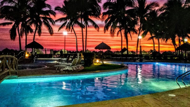 A swimming pool with lounge chairs and umbrellas, surrounded by palm trees, during sunset.