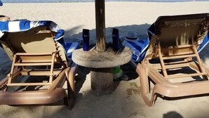 A beach scene with two sun loungers under a thatched umbrella, a small table with drinks, and a person in the distance.