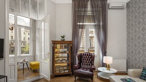 Et værelse med trægulv, en læderlænestol, et lille rundt bord med en lampe, en sofa med et plaide, et glasvitrineskab med bøger, og udsigt til bygninger gennem en åben dør.