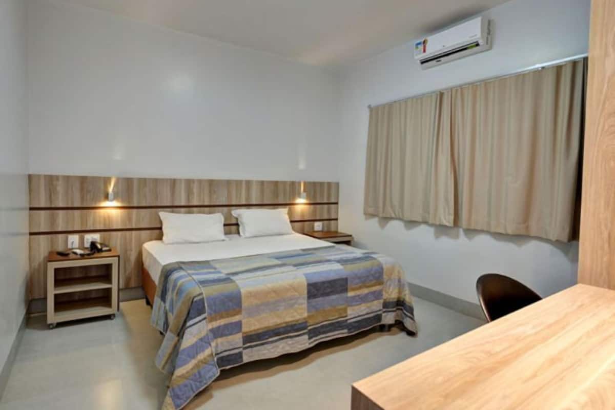Een hotelkamer met een bed, nachtkastjes, een bureau, een airconditioner en een raam met gordijnen.