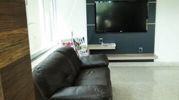 A modern living room with a leather sofa, a flat-screen TV mounted on the wall, and a window with magazines on the windowsill.