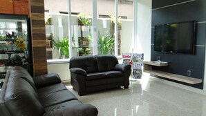 A modern living room with a black leather sofa, a flat-screen TV mounted on the wall, and a large window with plants visible outside. - Nacional Hotel Jataí (Jatai)