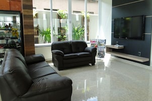 Uma sala de estar moderna com um sofá de couro preto, uma TV de tela plana fixada na parede e uma grande janela com plantas visíveis do lado de fora.