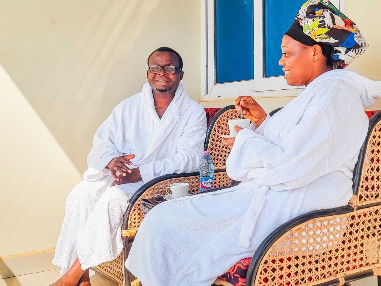 Two people in white robes sitting on wicker chairs with a water bottle and a cup on the table.