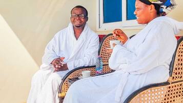 Two people in white robes sitting on wicker chairs with a water bottle and a cup on the table.