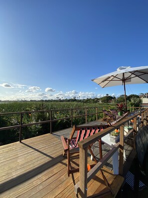 A wooden deck with lounge chairs and umbrellas, overlooking a lush green field and a clear blue sky.