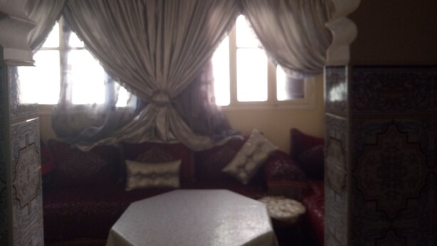 A room with a large window, a chandelier, and a sofa with multiple cushions. - Chez Simo un petit coin de relax (Safi)