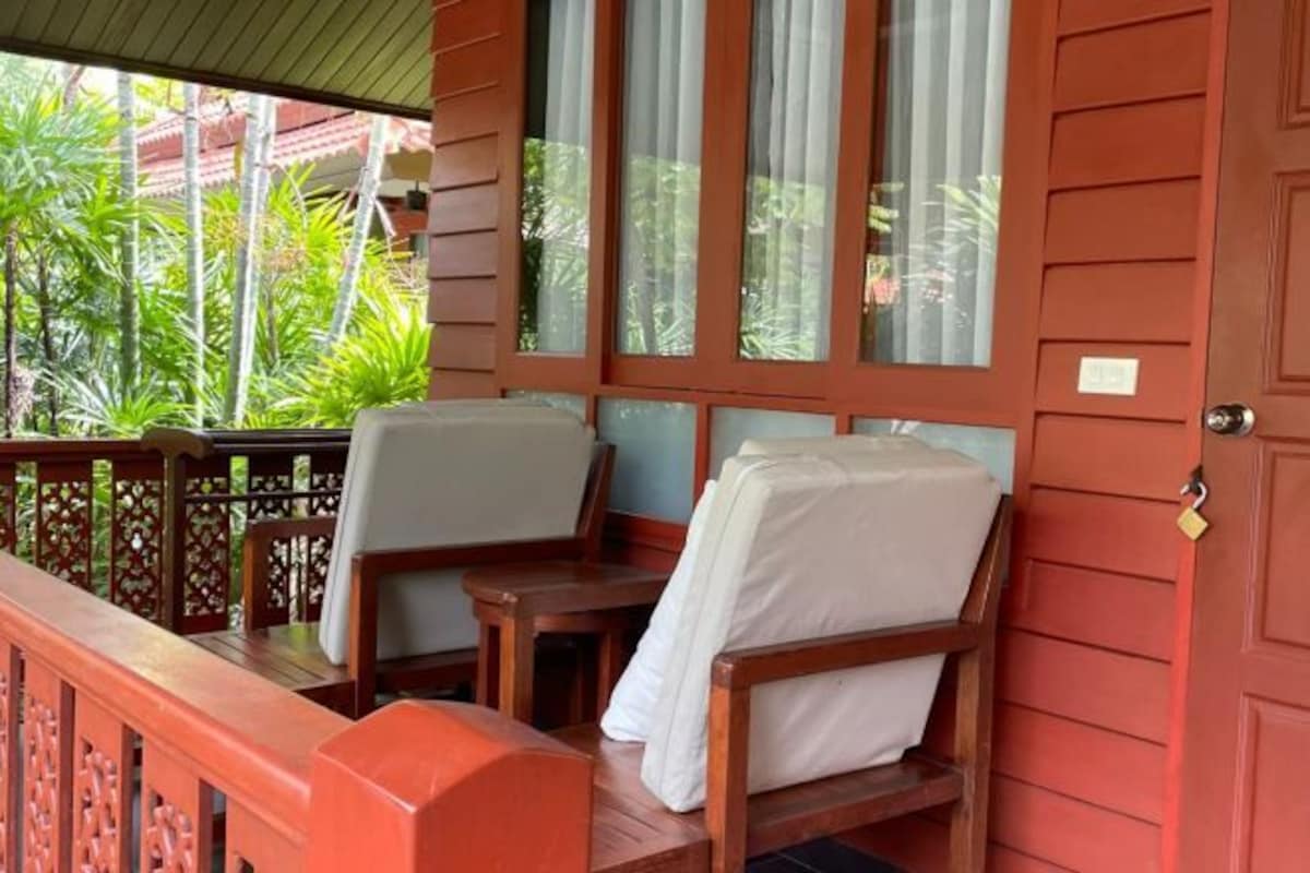 Una terraza de madera con una mesa y dos sillas, una puerta roja y vistas al verdor.