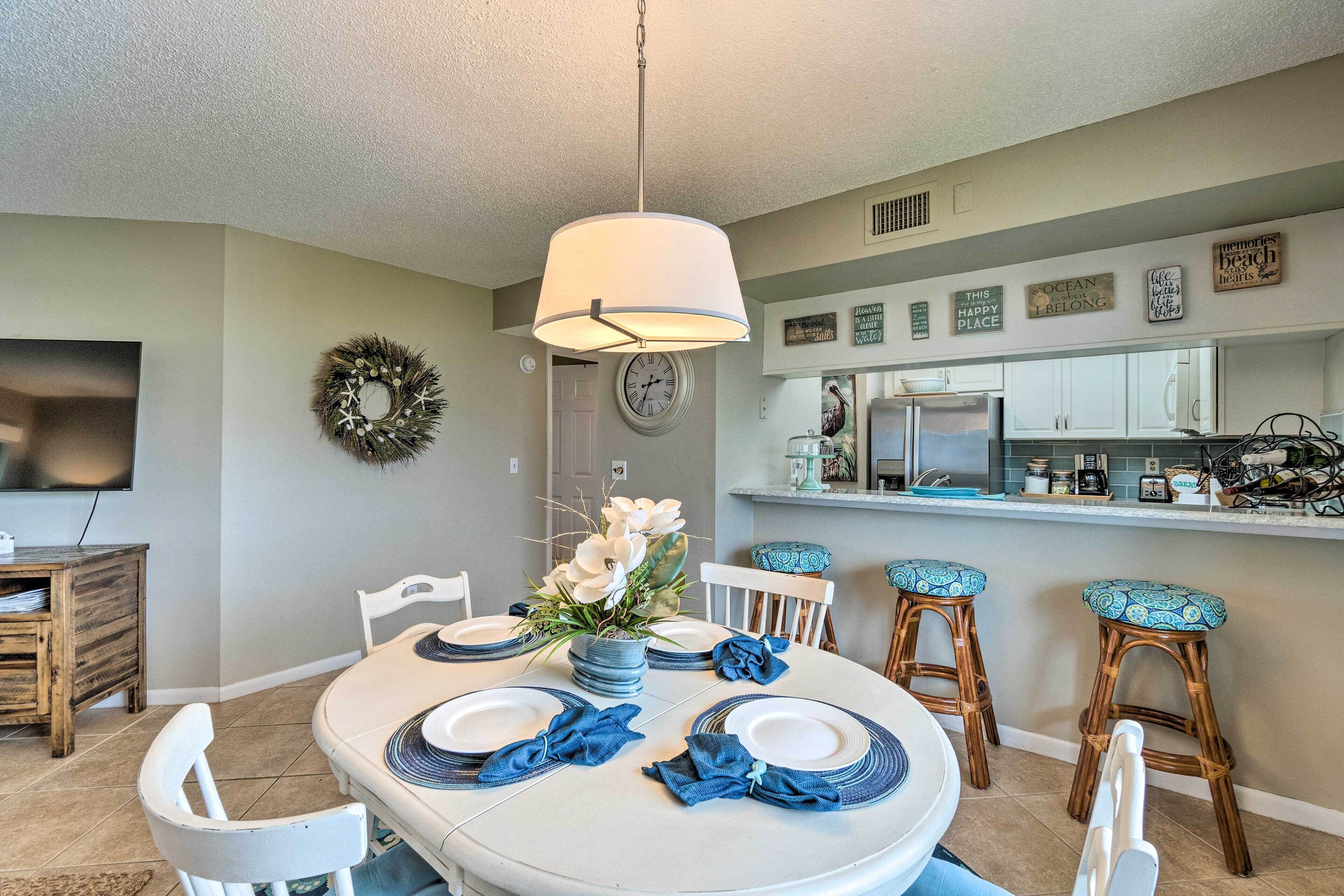 A dining area with a round table set for four, white chairs, a hanging light fixture, and a kitchen in the background.