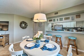 A dining area with a round table set for four, white chairs, a hanging light fixture, and a kitchen in the background. - Resort Condo w/ Beach Access on Anna Maria Island! (Bradenton Beach)