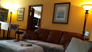 A room with a brown leather sofa, a small table, and a lamp. There are framed pictures on the wall. - A Clean, Safe, House full of steel balls & vintage game consoles (Richmond)