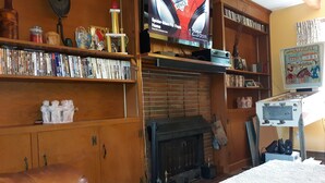 A cozy living room with a fireplace, a television mounted on the wall, and a bookshelf filled with books and decorative items. - A Clean, Safe, House full of steel balls & vintage game consoles (Richmond)
