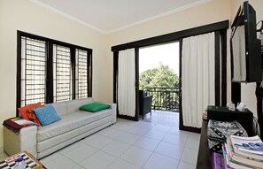 Un salon avec un canapé, une petite table et un téléviseur. Il y a des fenêtres avec des rideaux et un balcon d’où l’on peut admirer la vue sur les arbres.