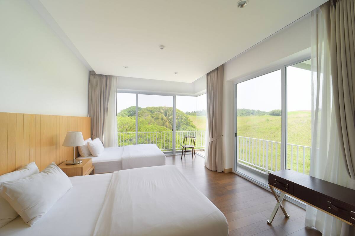 Habitación de hotel con dos camas, un escritorio y grandes ventanales con vistas a zonas verdes.