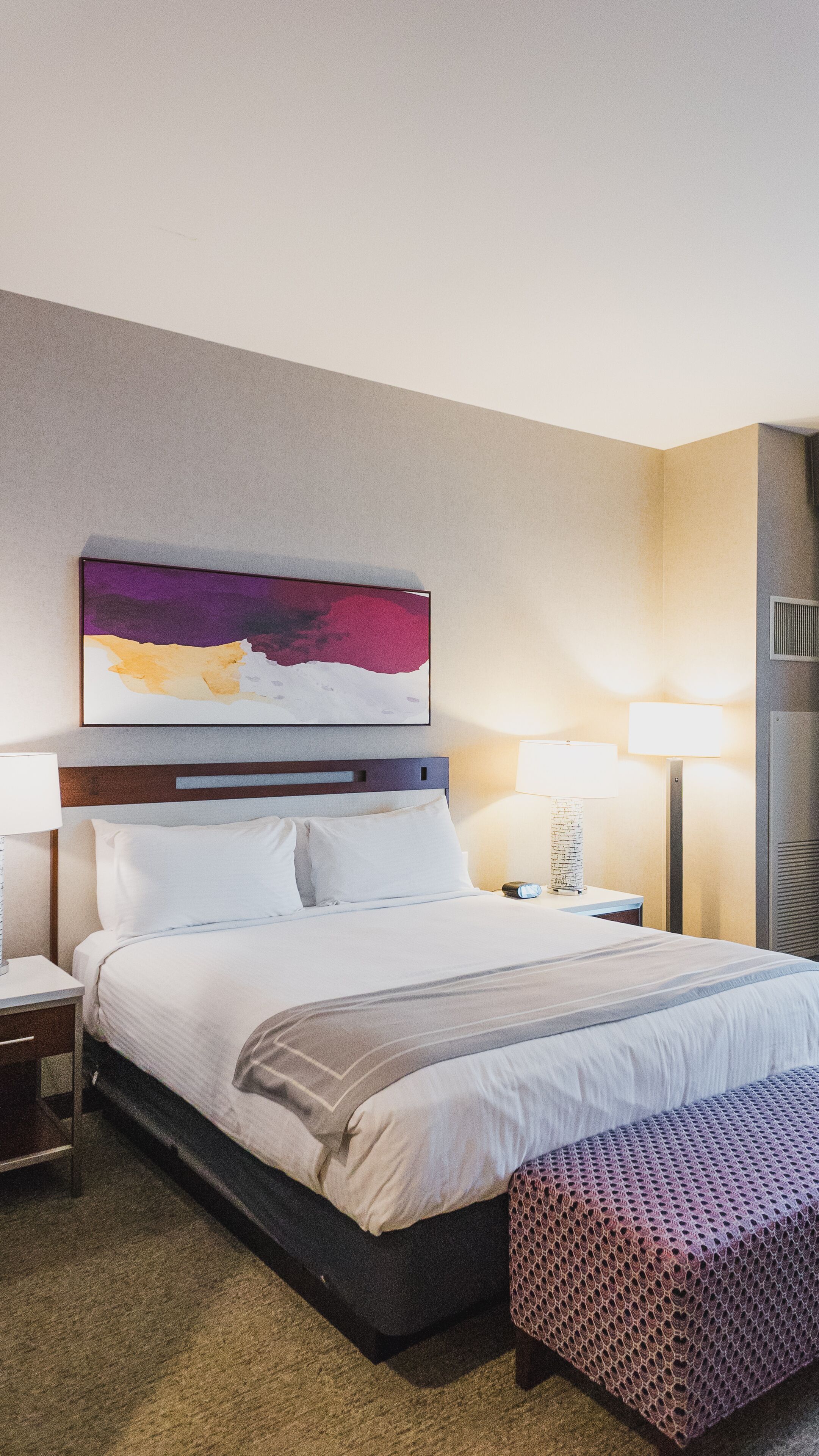 Ein Hotelzimmer mit einem großen Bett, einem Nachttisch, einer Lampe, einem Wandbild und einem gemusterten Hocker.