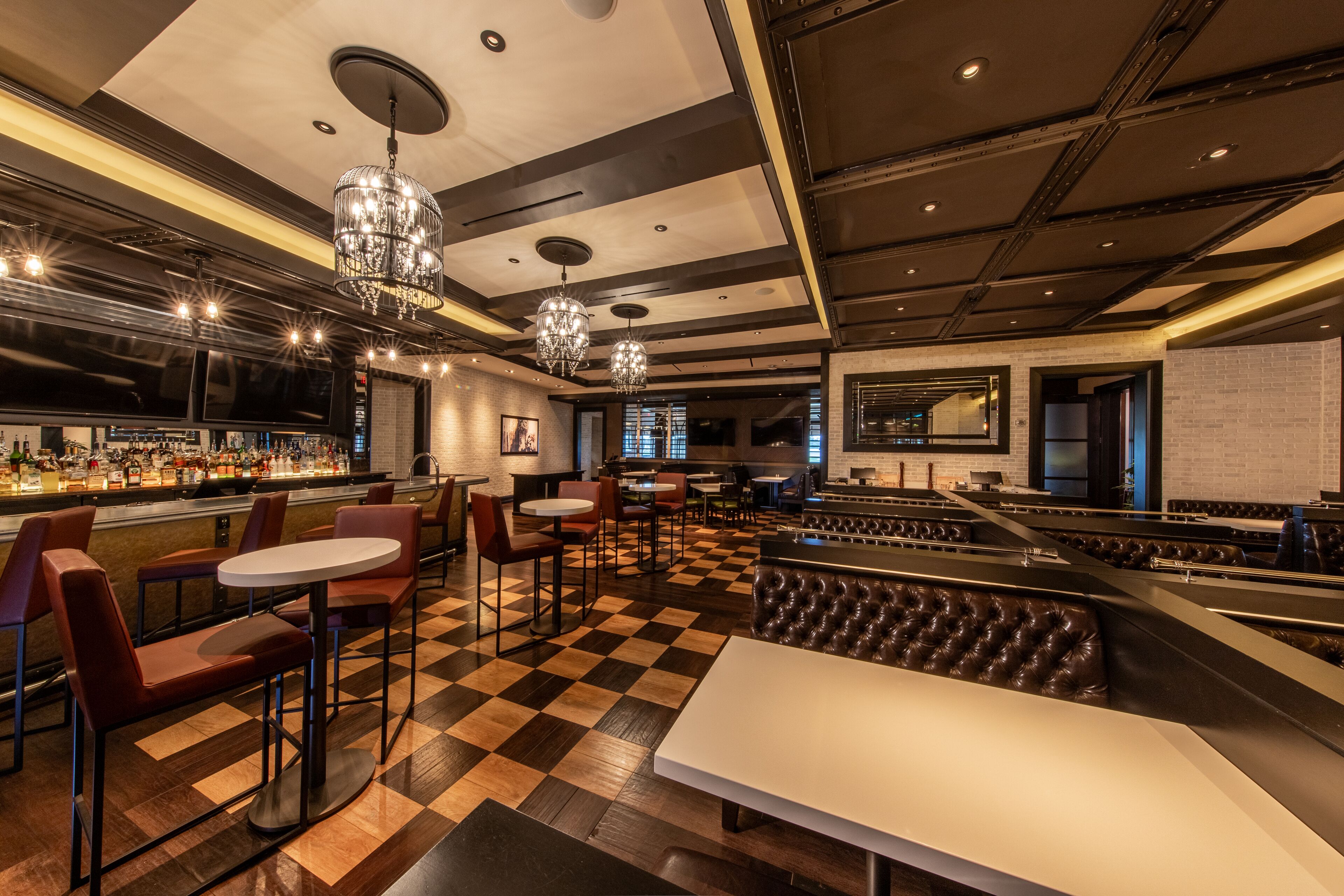 A restaurant with a checkered floor, bar area, and seating with a high ceiling and chandeliers.