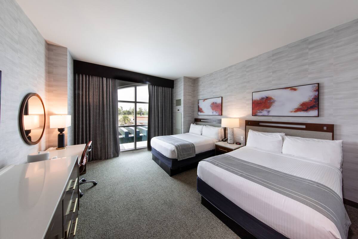 A hotel room with two beds, a desk, a mirror, and a large window with curtains.