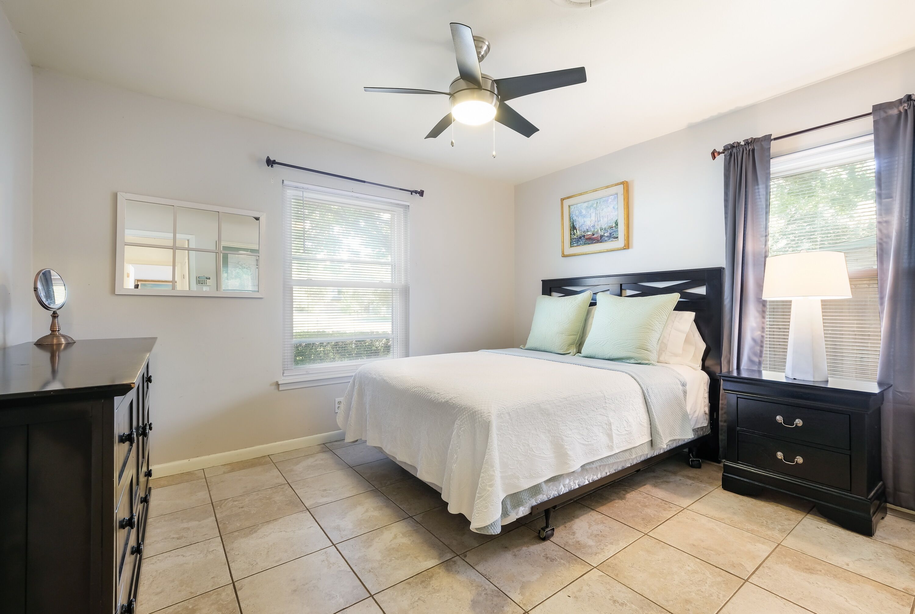 A bedroom with a bed, nightstand, ceiling fan, windows, and a dresser.
