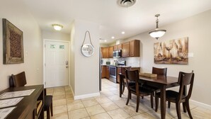 A dining area with a wooden table and chairs, a kitchen in the background, and a wall clock.
