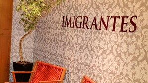Zona de recepción con una pared que muestra la palabra 'IMIGRANTES' y dos sillas rojas con una planta en maceta.
