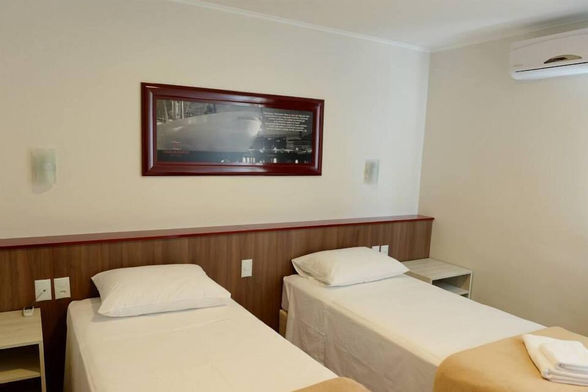 A hotel room with two single beds, a wooden headboard, a framed picture on the wall, and an air conditioning unit.