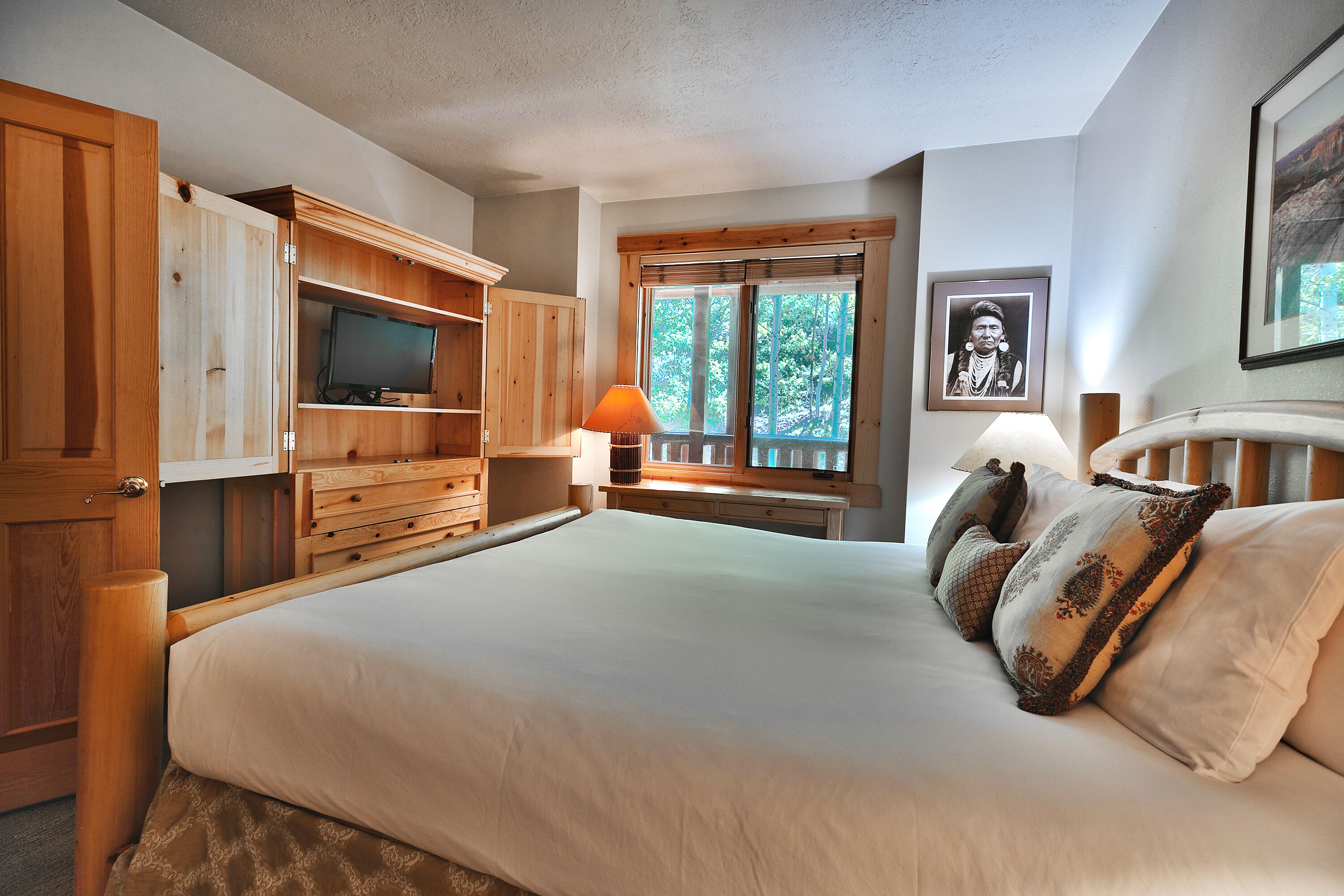 Un dormitorio con cama, armario de madera, televisor, ventana y un cuadro enmarcado.