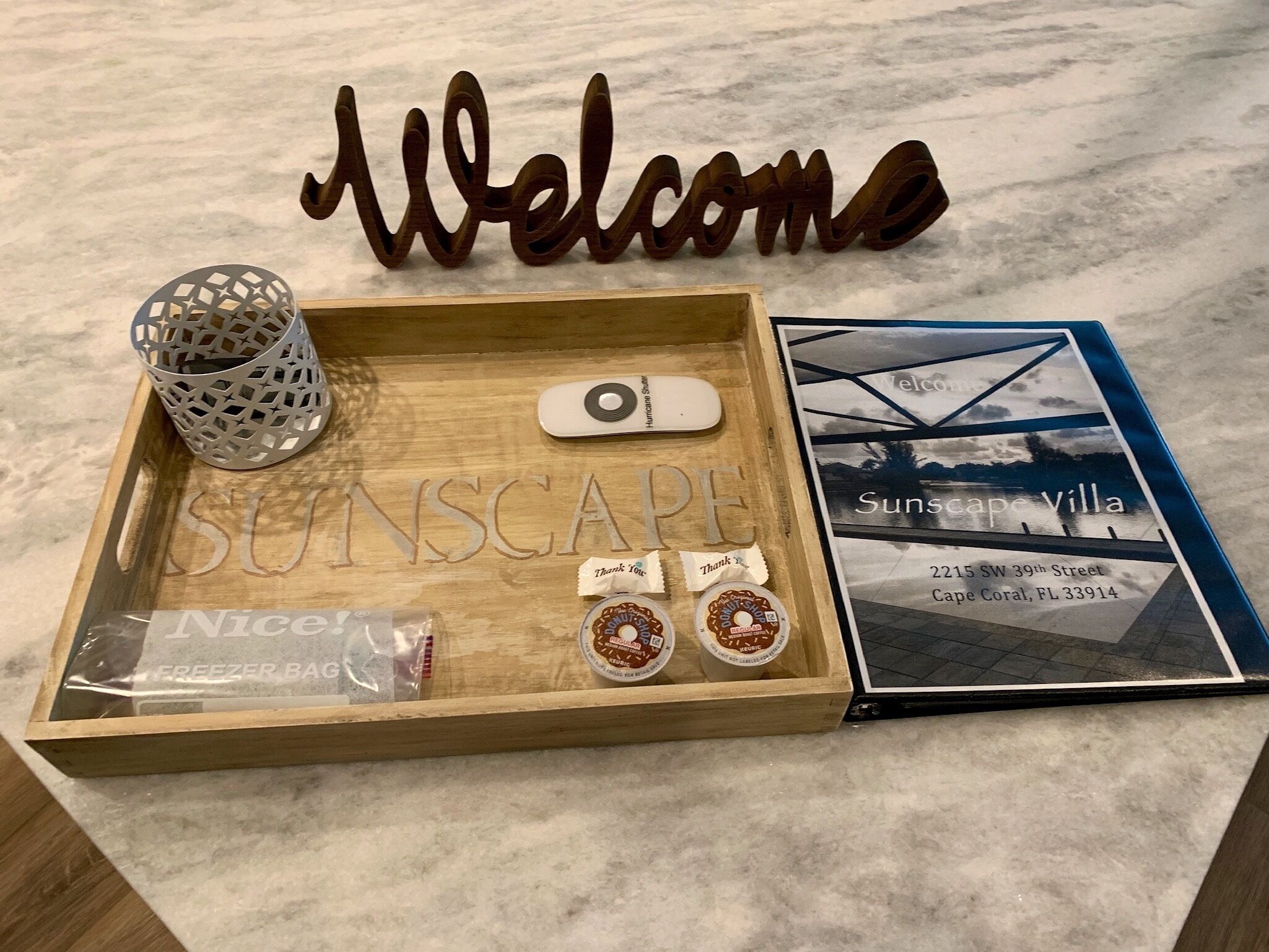 A wooden tray with a 'Welcome' sign, a Hurricane Shutter, a 'Sunscape Villa' brochure, and two 'Nice!' branded items.