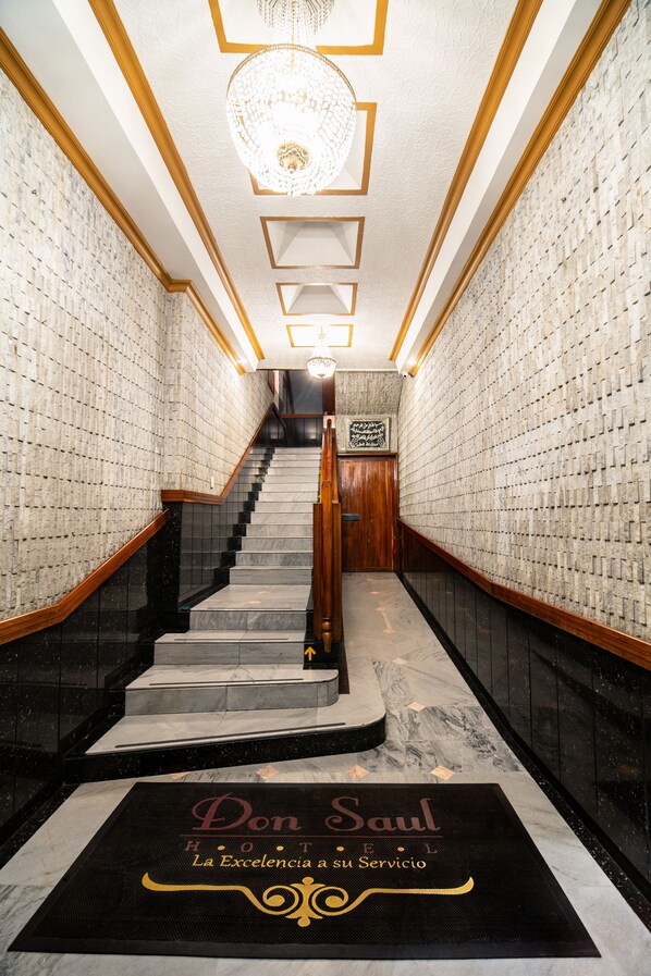 Interior de un hotel con una escalera de mármol, un candelabro y un letrero que dice 'Don Saul Hotel'.