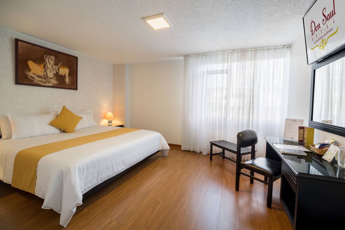 Una habitación de hotel con cama, escritorio, silla, lámpara y ventana con cortinas.