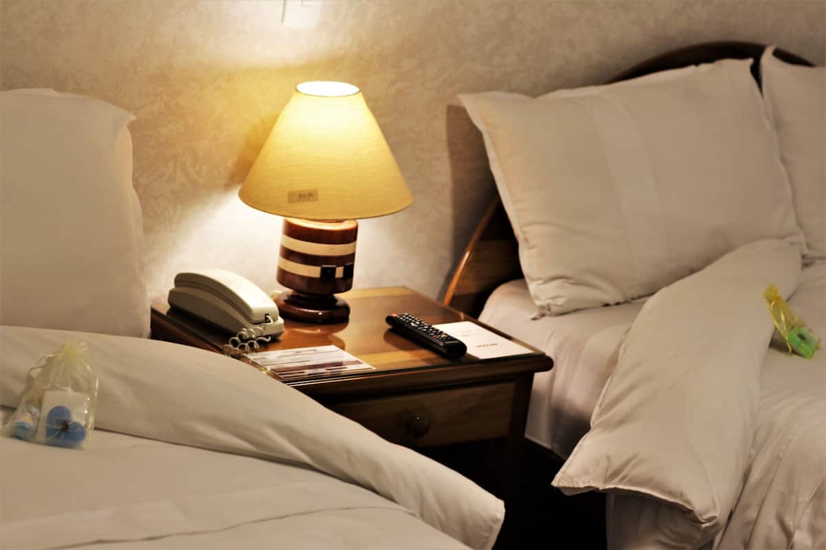 Una cama bien hecha con sábanas blancas, una lámpara de noche, un teléfono y un control remoto sobre una mesita de madera.