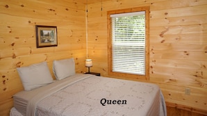 A wooden cabin bedroom with a queen-sized bed, a ceiling fan, a window with blinds, and a framed picture on the wall. - Cabin in the Woods-Gas Fireplace - Charcoal Grill - Pet Friendly (Whittier)
