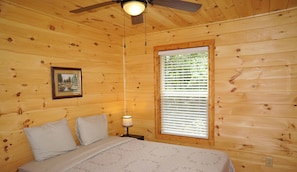 A wooden cabin bedroom with a queen-sized bed, a ceiling fan, a window with blinds, and a framed picture on the wall. - Cabin in the Woods-Gas Fireplace - Charcoal Grill - Pet Friendly (Whittier)
