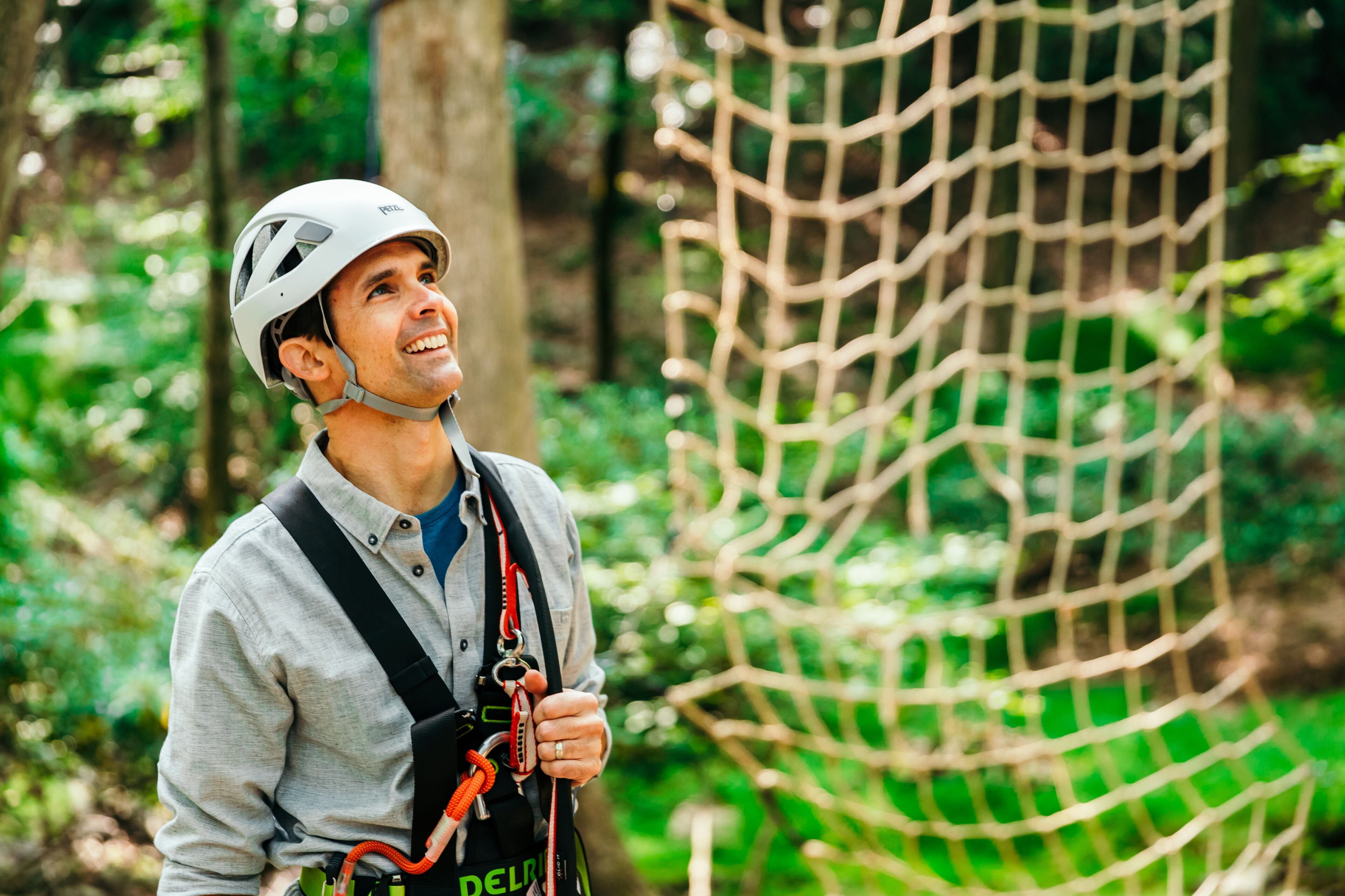 A person in a helmet and harness, standing next to a netted climbing structure in a forested area.