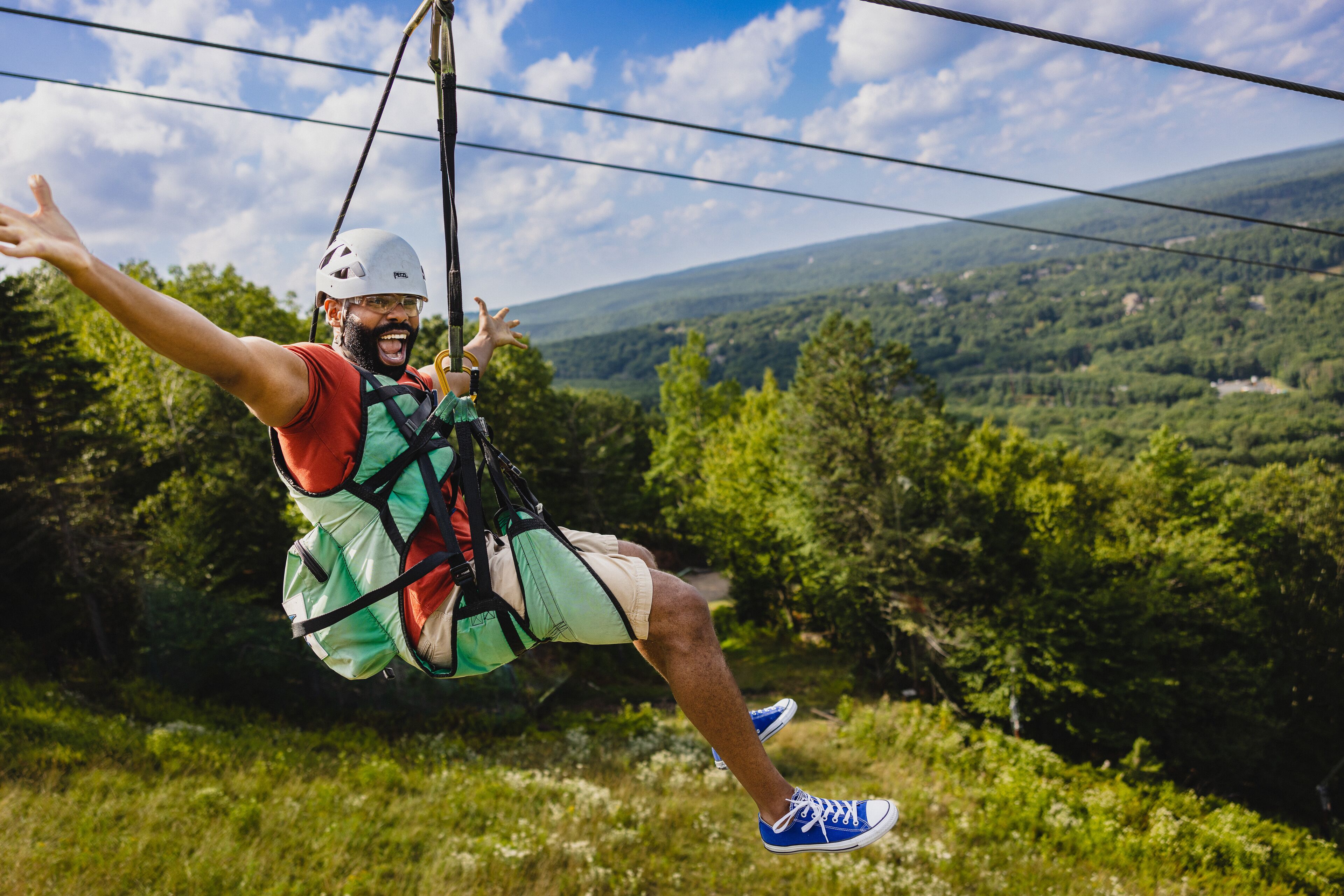 Een persoon op een zipline, vastgegespt met een harnas, draagt een helm, korte broek en sneakers.