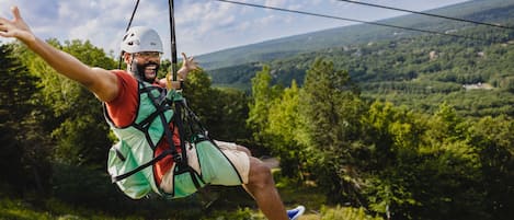 Een persoon op een zipline, vastgegespt met een harnas, draagt een helm, korte broek en sneakers.