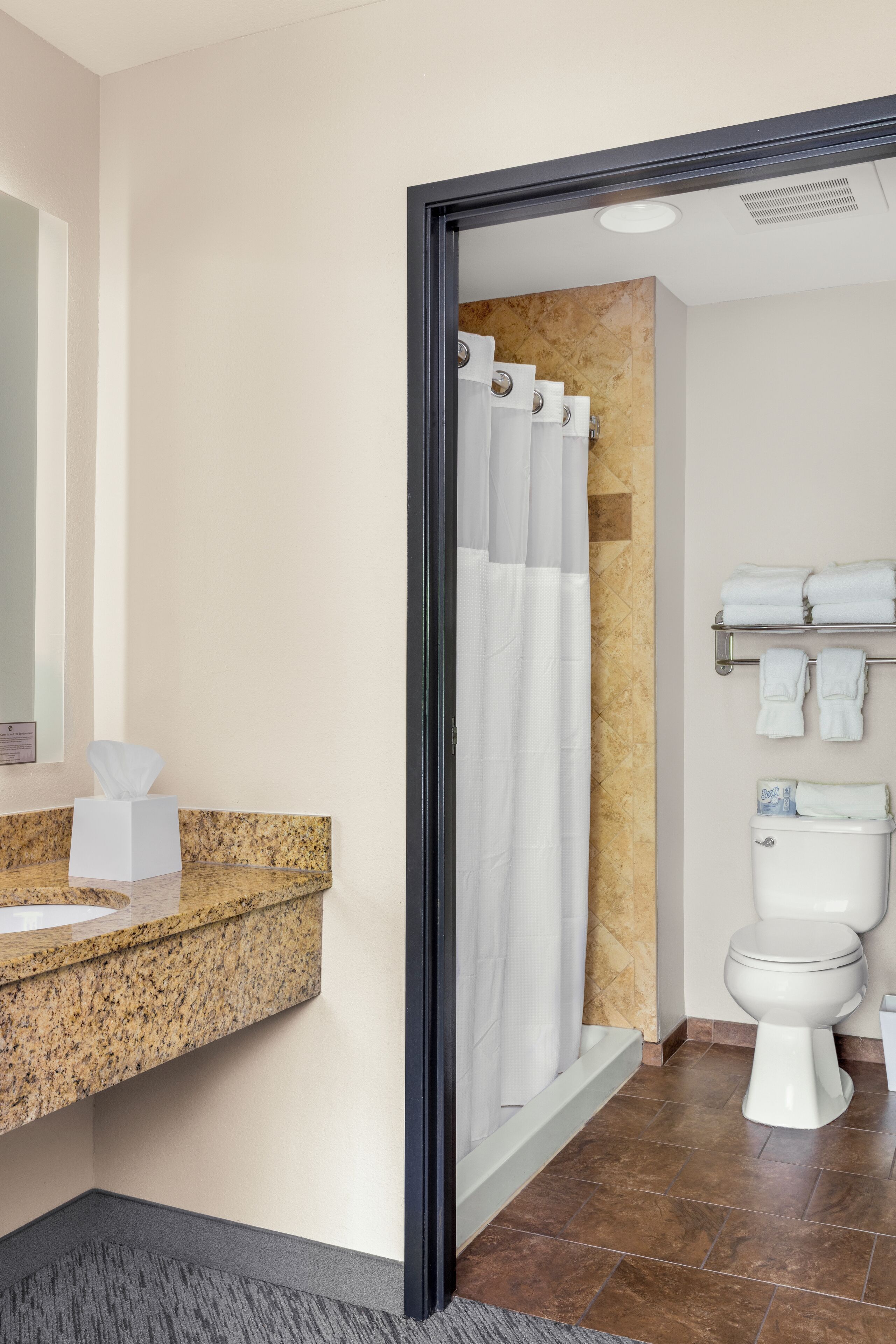 A modern bathroom with a granite countertop, a white toilet, a shower curtain, and a towel rack with folded towels.